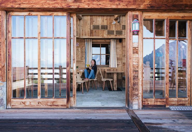 Woman reading on the wooden terrace of a chalet, seen through large glass doors.
