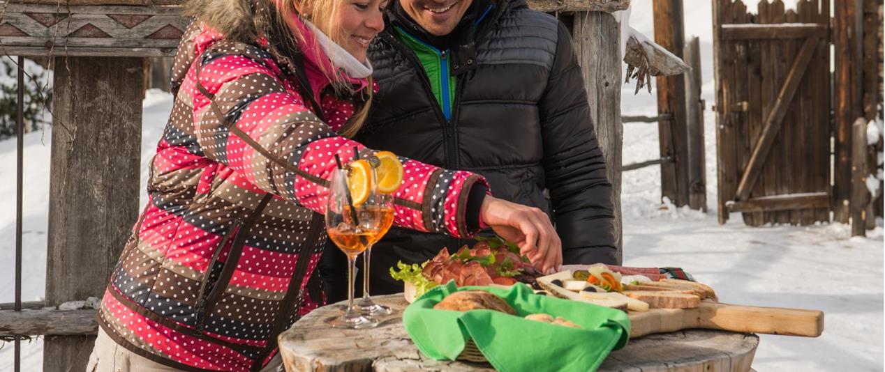Due persone durante un aperitivo sulla neve davanti alla Graziani Lodge con sculture in legno e bevande.