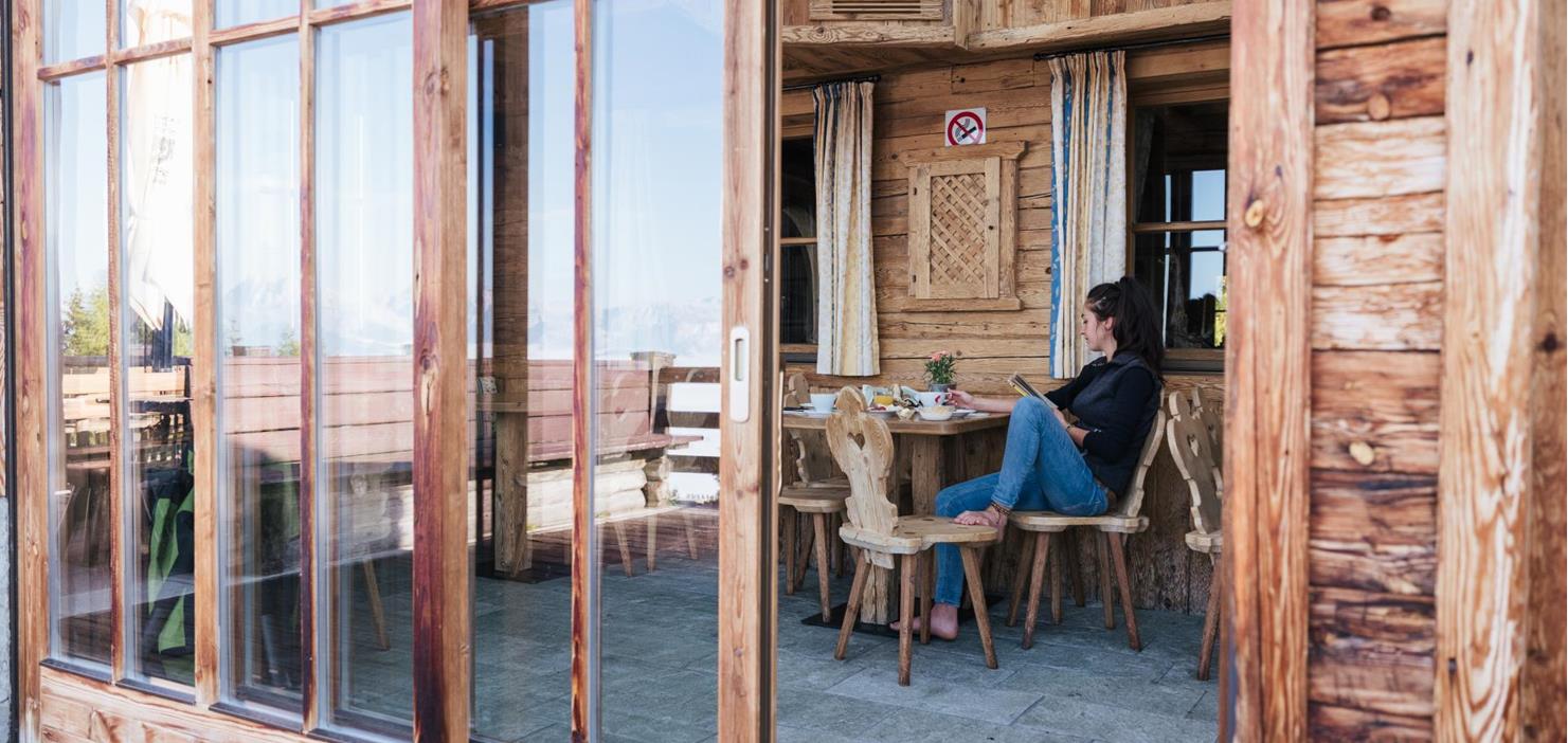 Frau beim Frühstück auf der Holzterrasse eines Chalets mit Blick durch große Glastüren.