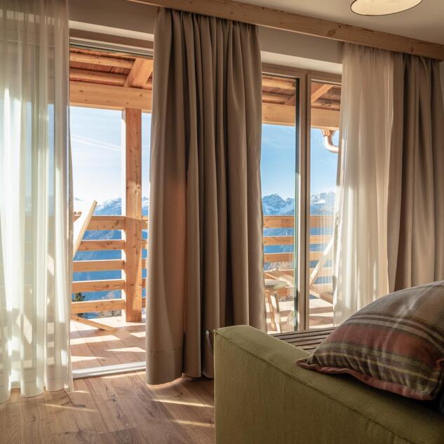 Blick durch die Fensterfront auf den Holzbalkon eines Chalets mit Bergpanorama.