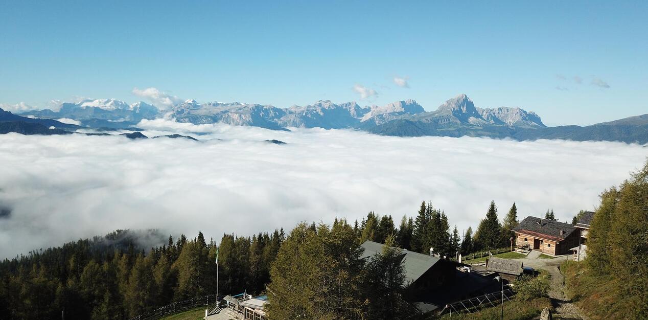 Blick auf Chalets und Nadelwald oberhalb eines Wolkenmeers und den Dolomiten im Hintergrund.