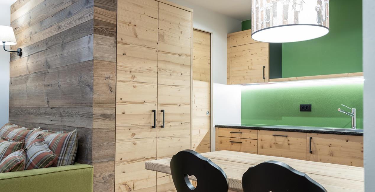 Zona giorno e pranzo della Ciasa Rosina con divano verde, tavolo in legno e angolo cottura in legno chiaro