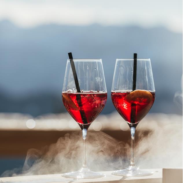 Two glasses of red cocktails on a sunny terrace, with a blurred mountain panorama in the background.