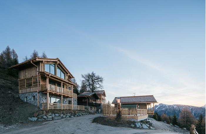 Wooden chalets and coniferous trees under a blue sky with views of the Dolomites.