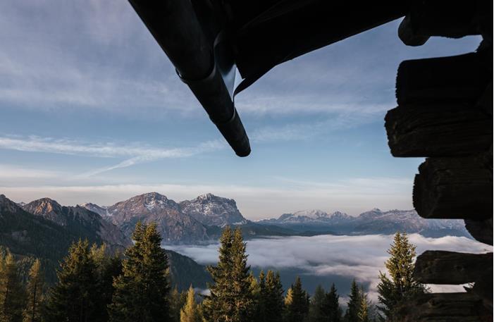 Ausblick auf die Dolomiten mit Nadelwald, Nebel im Tal und Holzhütte im Vordergrund.