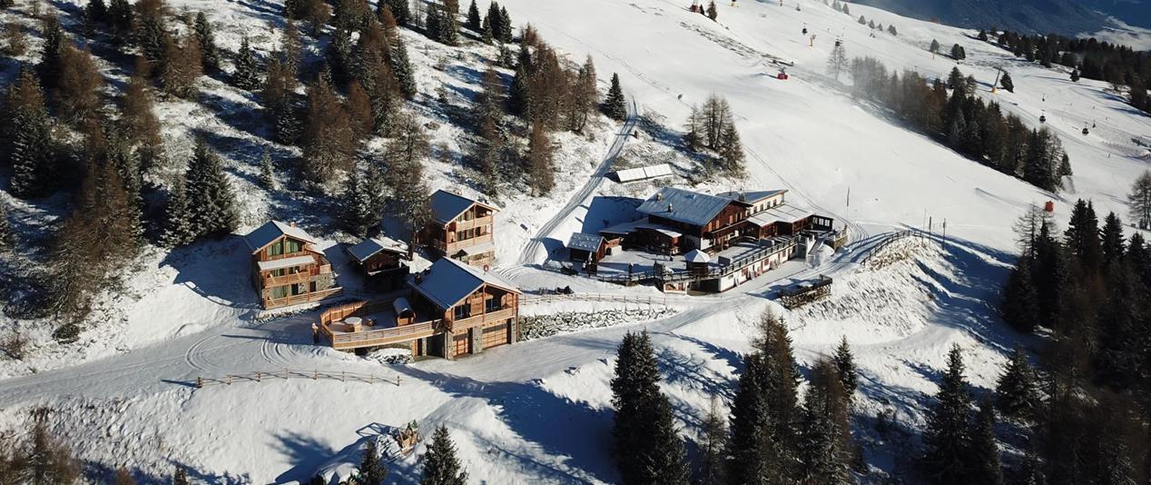 Luftaufnahme der Graziani Hütte und umliegender Holzchalets inmitten einer verschneiten Skilandschaft.