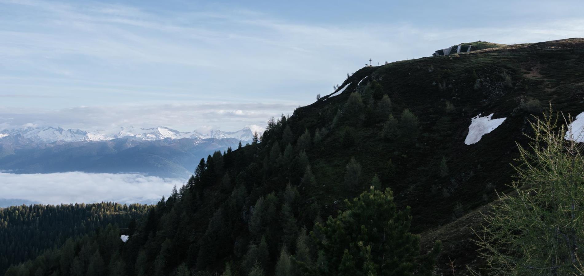 Bewaldeter Berghang am Kronplatz mit Schneefeldern und Blick auf entfernte Gipfel.