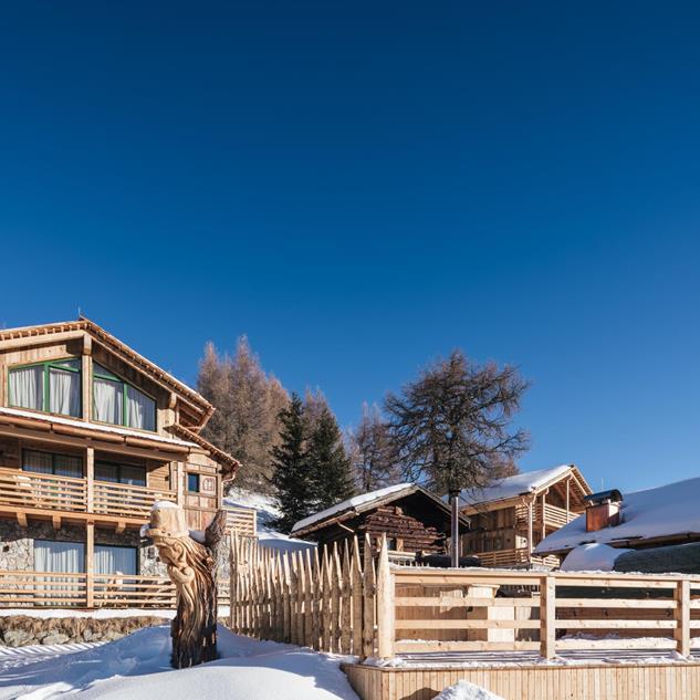 Wooden chalets with balconies and snow-covered roofs in a winter landscape under a blue sky.
