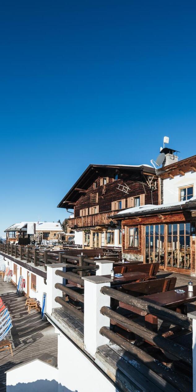 Sunny wooden terrace of the Graziani mountain hut with tables and benches, surrounded by a snow-covered winter landscape.
