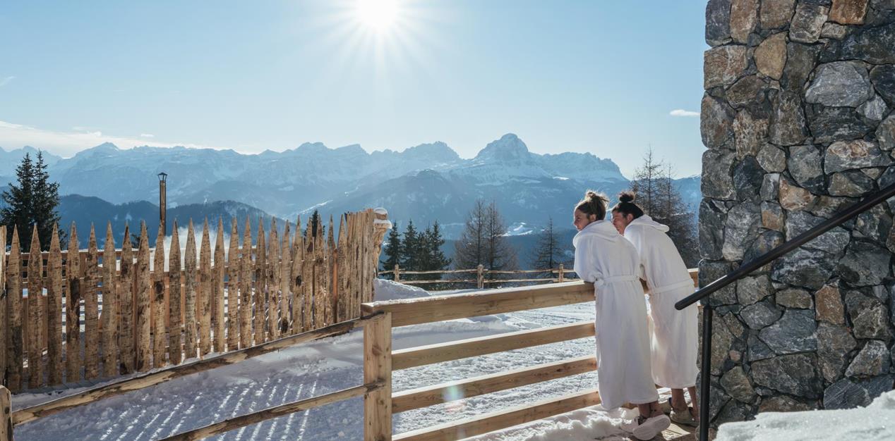 Paar in weißen Bademänteln genießt den Blick auf die verschneite Berglandschaft bei strahlendem Sonnenschein.