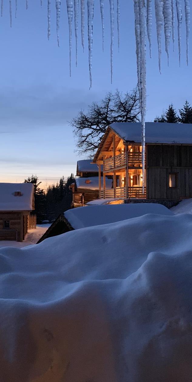Snow-covered wooden chalets with warmly lit windows at dusk and long icicles in the foreground.