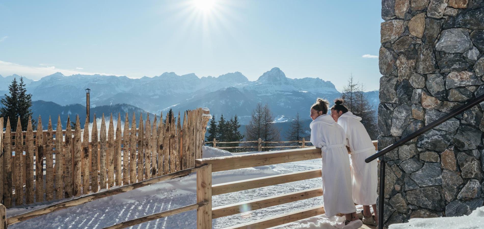 Ein Paar in weißen Bademänteln angelehnt an einem Holzzaun mit Blick in die winterliche Berglandschaft.